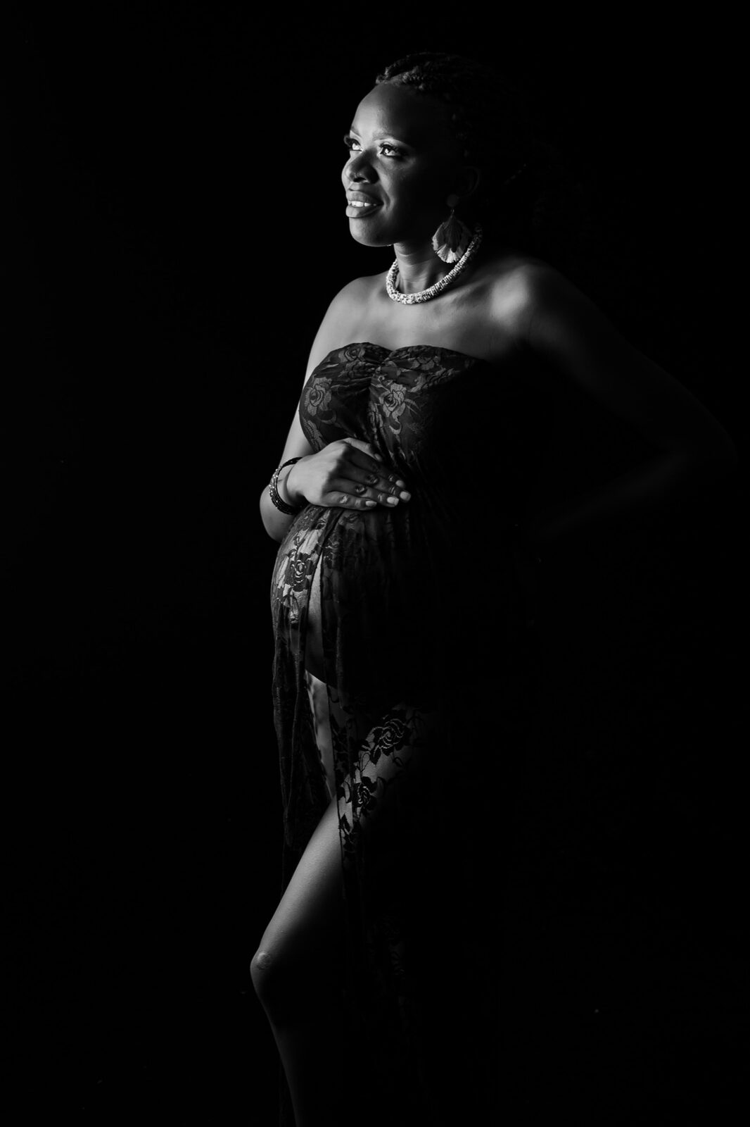 Smiling pregnant woman in a strapless lace gown cradling her belly, lit dramatically against a black backdrop in a black and…